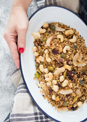 Woman hand holding a metallic white tray full of delicious granola over a rustic napkin on a white and grey background.