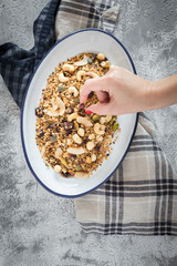 Woman hand taking a pistachio from a metallic white tray full of delicious granola over a rustic napkin on a white and grey background.