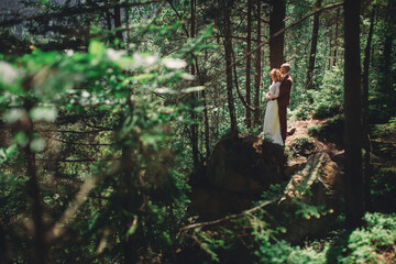 Happy stylish couple newlyweds in the green forest on summer day. bride in long white dress and groom in red suit are hugging. wedding day.