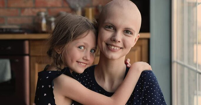 Young bald sick mother cancer patient embrace little daughter in kitchen smile look at camera. Caring kid express tender, gives support, feeling devotion to parent with oncology disease. Love concept