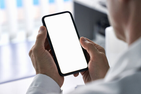 Male doctor holding cell phone in hands using blank white mockup screen technology ehealth mobile app for medical healthcare telemedicine ads, e telehealth online applications. Over shoulder view