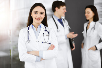 Fototapeta premium Smiling intelligent woman-doctor is standing with arms crossed in sunny clinic together with her colleagues at the background. Perfect medical service in a hospital