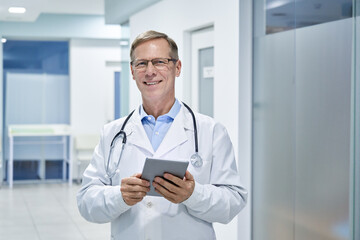 Smiling old middle aged doctor holding using digital tablet standing in hospital looking at camera. Online healthcare telemedicine ehealth services,medical internet technology apps,telehealth concept.