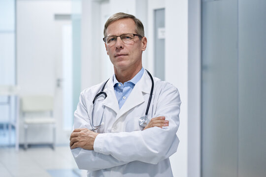 Trustworthy proud mature old medical professional doctor stands arm crossed in hospital. Middle aged male senior physician practitioner wears white coat, stethoscope, glasses looks at camera. Portrait