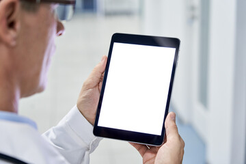 Male doctor holding digital tablet in hands using blank white mockup screen technology for medical healthcare tech website ads concept, video call e telehealth online appointment. Over shoulder view