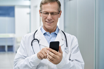 Old male doctor holding smartphone using mobile phone e health technology in hospital. Modern tech medical healthcare patients virtual consultation app, telehealth online, tele medicine appointment.