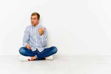 Young caucasian man sitting on the floor points with thumb finger away, laughing and carefree.