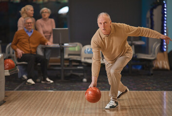 Full length portrait of active senior man playing bowling and throwing ball by lane with group of...
