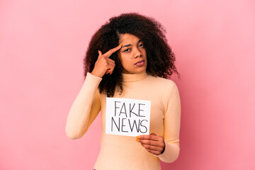 Young african american curly woman holding a fake news on a placard pointing temple with finger, thinking, focused on a task.