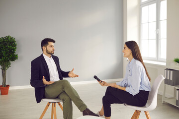 Young man sitting on chair in television studio and giving interview to journalist. Happy smiling TV host with microphone talking to famous person and asking questions about his opinion and experience