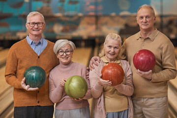 Waist up portrait of group of happy senior people holding bowling balls and smiling at camera while enjoying active entertainment at bowling alley, copy space