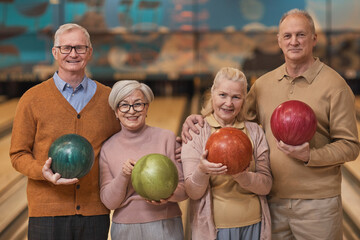 Fototapeta premium Waist up portrait of group of happy senior people holding bowling balls and smiling at camera while enjoying active entertainment at bowling alley, copy space