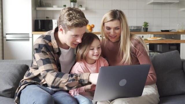Happy family young parents and kid child daughter using laptop computer having fun sitting on sofa at home, enjoying talking, doing e commerce shopping online, choosing buying on website together.
