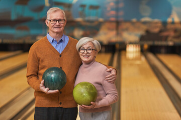 Waist up portrait of happy senior couple holding bowling balls and smiling at camera while enjoying...