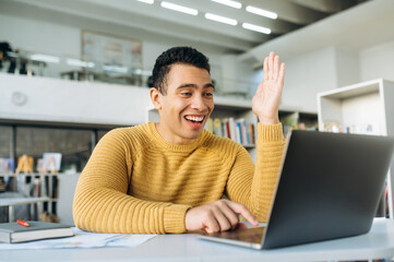 Handsome young adult man at online lesson or webinar training. Hispanic male student or employee sitting at the desk, using laptop and smiling. Studying remotely