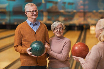 Fototapeta premium Waist up portrait of group of happy senior people holding bowling balls and chatting while enjoying active entertainment at bowling alley, copy space