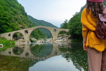 White girl backpacker looking medieval old ottoman stone structure with reflect on the water. Devil Bridge in Bulgaria with legend. Lonely woman on a trip in a river.