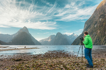 Tourist travel photographer taking pictures with professional camera on tripod on adventure travel vacation in mountain landscape. Milford Sound, Fiordland National Park, South Island, New Zealand. - obrazy, fototapety, plakaty