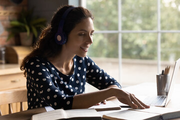 Happy millennial Caucasian woman in headphone sit at table at home study online on computer. Smiling young female student take distant course or web training on laptop. Education concept.