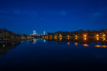 Fototapeta na wymiar Lake in park during blue hour with illuminated office buildings and apartment blocks