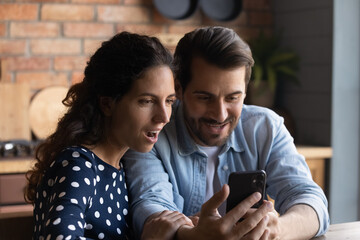 Surprised young Caucasian couple hold cellphone feel stunned with online promotion discount or sale offer. Amazed millennial man and woman look at smartphone screen read unbelievable news on web.