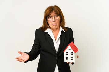 Middle aged real estate agent holding a house model isolated shrugs shoulders and open eyes...