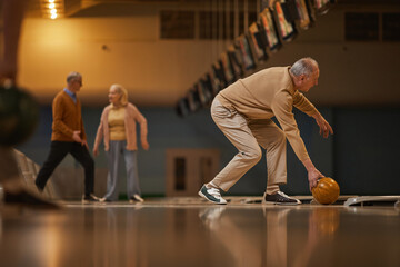 Wide angle side view at senior couple man playing bowling while enjoying active entertainment at bowling alley, copy space