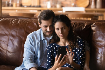 Young Caucasian man and woman sit on sofa at home use cellphone make self-portrait family picture together. Loving couple relax on couch talk on video call or take selfie on modern smartphone.