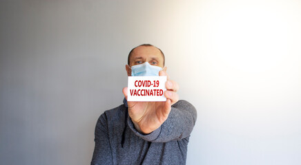 COVID-19 vaccinated symbol. White card with words 'COVID-19 vaccinated'. A young man in a grey wear and medical mask. Sunshine. Beautiful white background. COVID-19 vaccinated coronavirus concept.