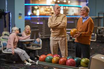 Fototapeta premium Portrait of two senior men playing bowling together with friends in background while enjoying active entertainment at bowling alley, copy space