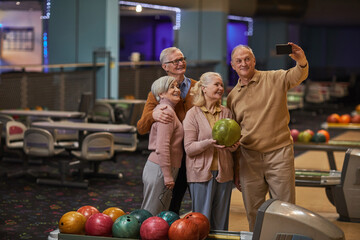 Fototapeta premium Wide angle view at group of senior people taking selfie photos playing bowling and enjoying active entertainment at bowling alley, copy space