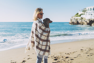 Charming blonde woman wearing blue jeans with a plaid on her shoulders standing with her dog in her arms on a blurred background of a sea shore. Student girl walking the beach side with her pup. - 408163748