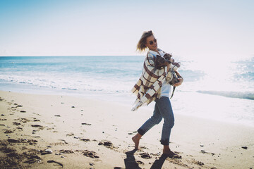 Smiling blonde woman in casual clothes with a blanket on her shoulders walking barefoot alongside the sea shore with her lovely dog in her arms. Woman spending sunny day with her pup on a sandy beach. - 408163754