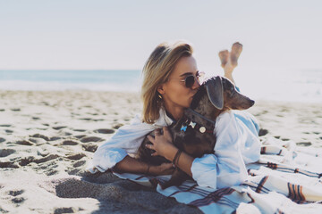 Fototapeta na wymiar Cheerful female in casual outfit laughing and kissing dog while lying on a sand beach at the day off. Young woman is looking at her cute puppy while relaxing on a warm sunny day outdoors.