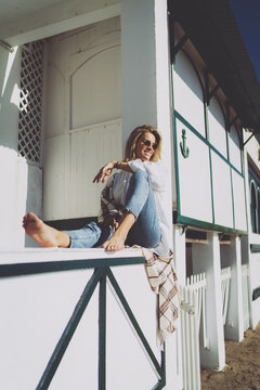 Cheerful blonde female in sunglasses sitting barefoot on a parapet of a beach house on a day off. Smiling caucasian female wearing white shirt and blue jeans enjoying warm sunny day on a beachside.