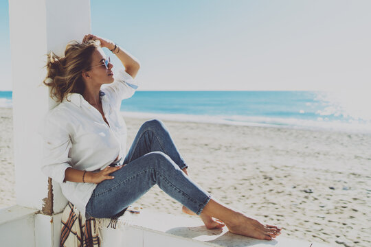 Thoughtful attractive woman dressed in stylish casual clothes enjoying good sunny weather on a sunny beach. Gorgeous model look blonde female in sunglasses relaxing on a warm windy day on a beachside.