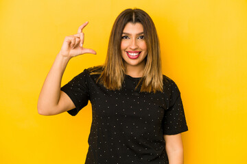 Young indian woman isolated on yellow background holding something little with forefingers, smiling and confident.