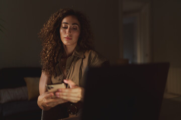 Beauty blogger female checking comments to the post by a smartphone. Attractive young woman holding a mobile phone while sitting behind a laptop at home. Student girl chatting online via social media