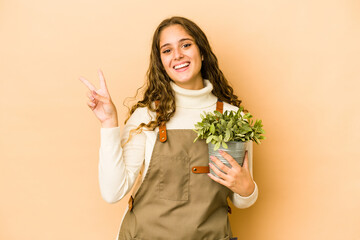 Young caucasian gardener woman holding a plant isolated joyful and carefree showing a peace symbol with fingers.