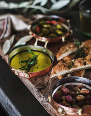 Pickled Greek olives, olive oil in copper jars and herbed focaccia slices on rustic wooden board, selective focus, close-up. Traditional Mediterranean meze appetizers platter