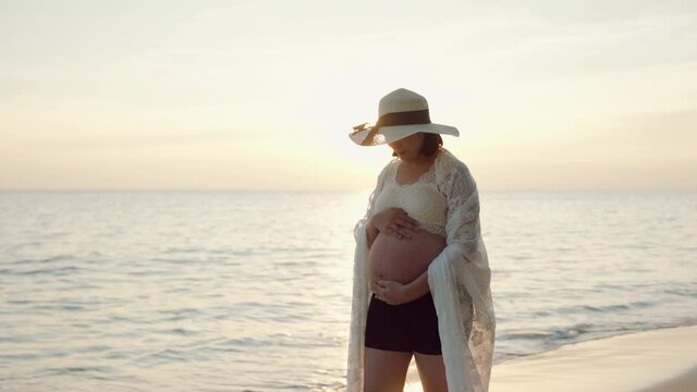 Young Asian pregnant woman walking and touch her belly expect for baby good health on the beach at sunset. concept of health, prenantcy and mother