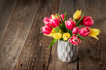 Fresh spring yellow and pink tulips bouquet in blue vase standing on rustic wooden table background. Festive flowers for mother's or women's day. Mockup for greeting square card. Copy space.