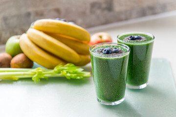 Two glasses of fresh green smoothie decorated with chia seeds and blueberry on the kitchen table with ingredients. Healthy food, detox eating, vegetarian, vegan diet. Selective focus, copy space.