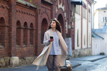 Beautiful brunette woman walking in a new city with a phone in her hands and a suitcase, a tourist...