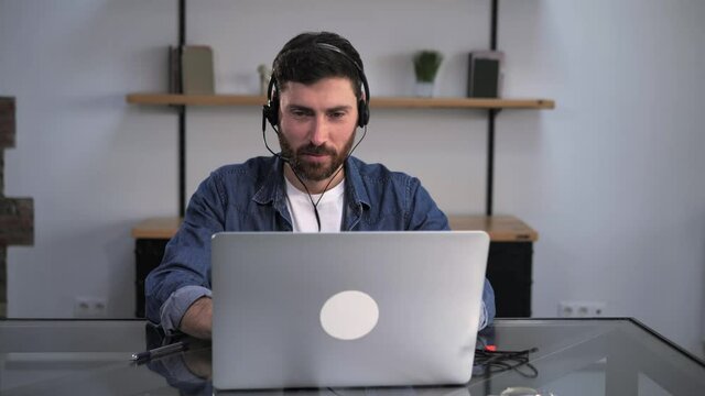 Young attractive bearded businessman working from home office using laptop headphones microphone as technical support agent. Call center worker receiving video call talking to customer smiling.
