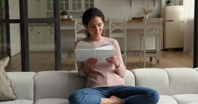 Relaxed millennial pretty woman sitting on comfortable couch with paper book in hands, involved in reading favorite novel bestseller literature, enjoying spending calm peaceful weekend at home.