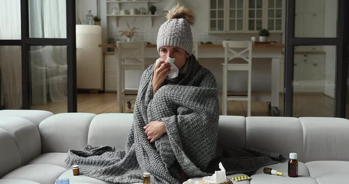 Unhealthy young woman wearing warm hat and covered in knitted plaid sneezing, wiping runny nose with paper tissue, feeling unwell, curing flu grippe disease with medical treatment alone at home.