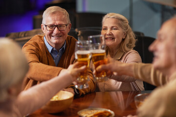 Portrait of smiling senior people drinking beer in bar and clinking glasses while enjoying night out with friends