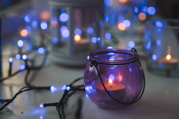 Fototapeta na wymiar Close-up of burning tea candles in lantern and glasses on sill. Blue garland light. Hygge christmas decorations.