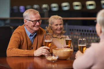 Portrait of modern senior couple drinking beer in bar and laughing while enjoying night out with friends, copy space
